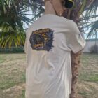 1 1 DOS - PredaColor Mannequin wearing a light shirt with a Predator Gator Liner graphic on the back, leaning against a palm tree outdoors, wearing a yellow-and-black cap.