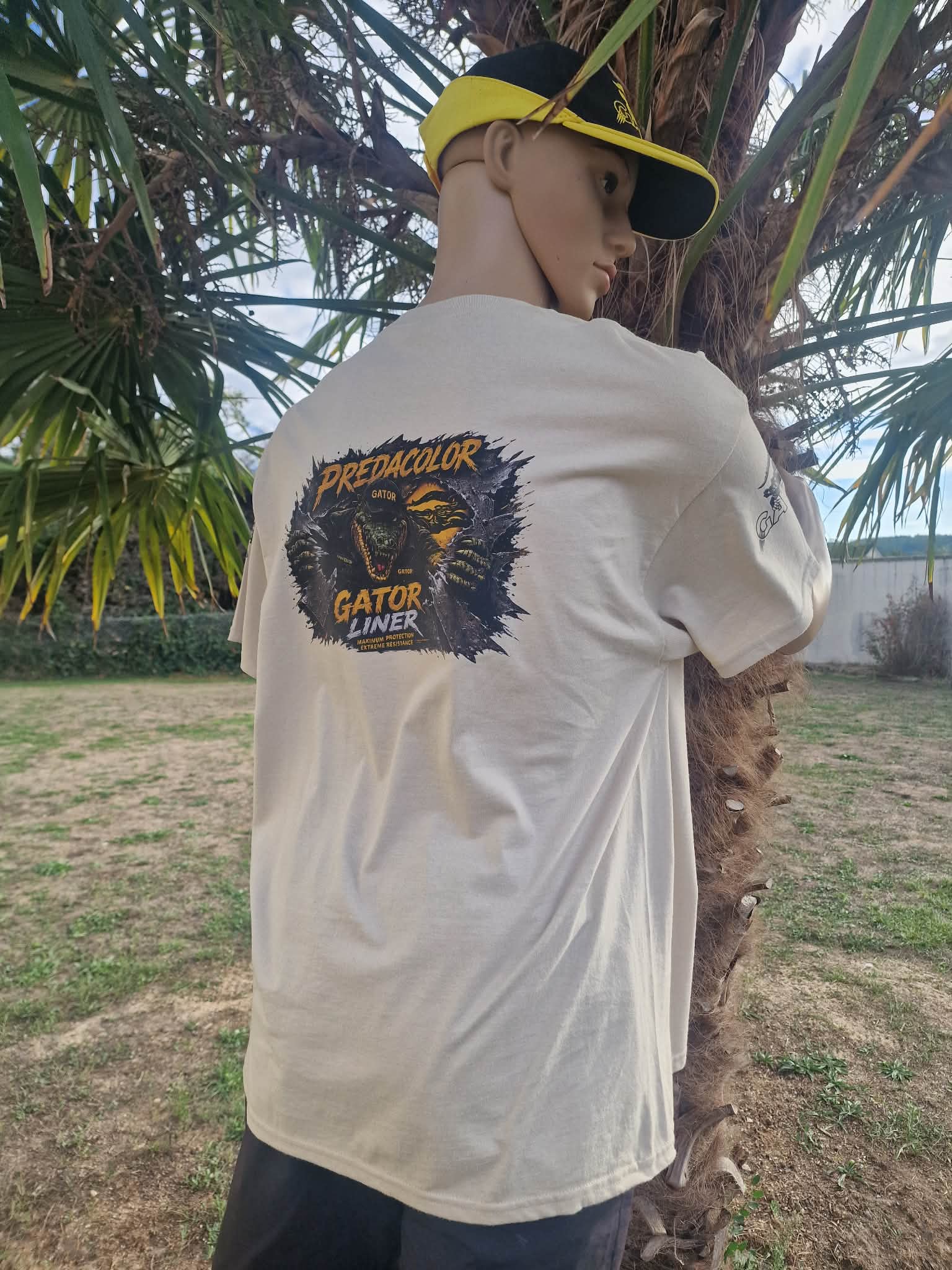 1 1 DOS - PredaColor Mannequin wearing a light shirt with a Predator Gator Liner graphic on the back, leaning against a palm tree outdoors, wearing a yellow-and-black cap.