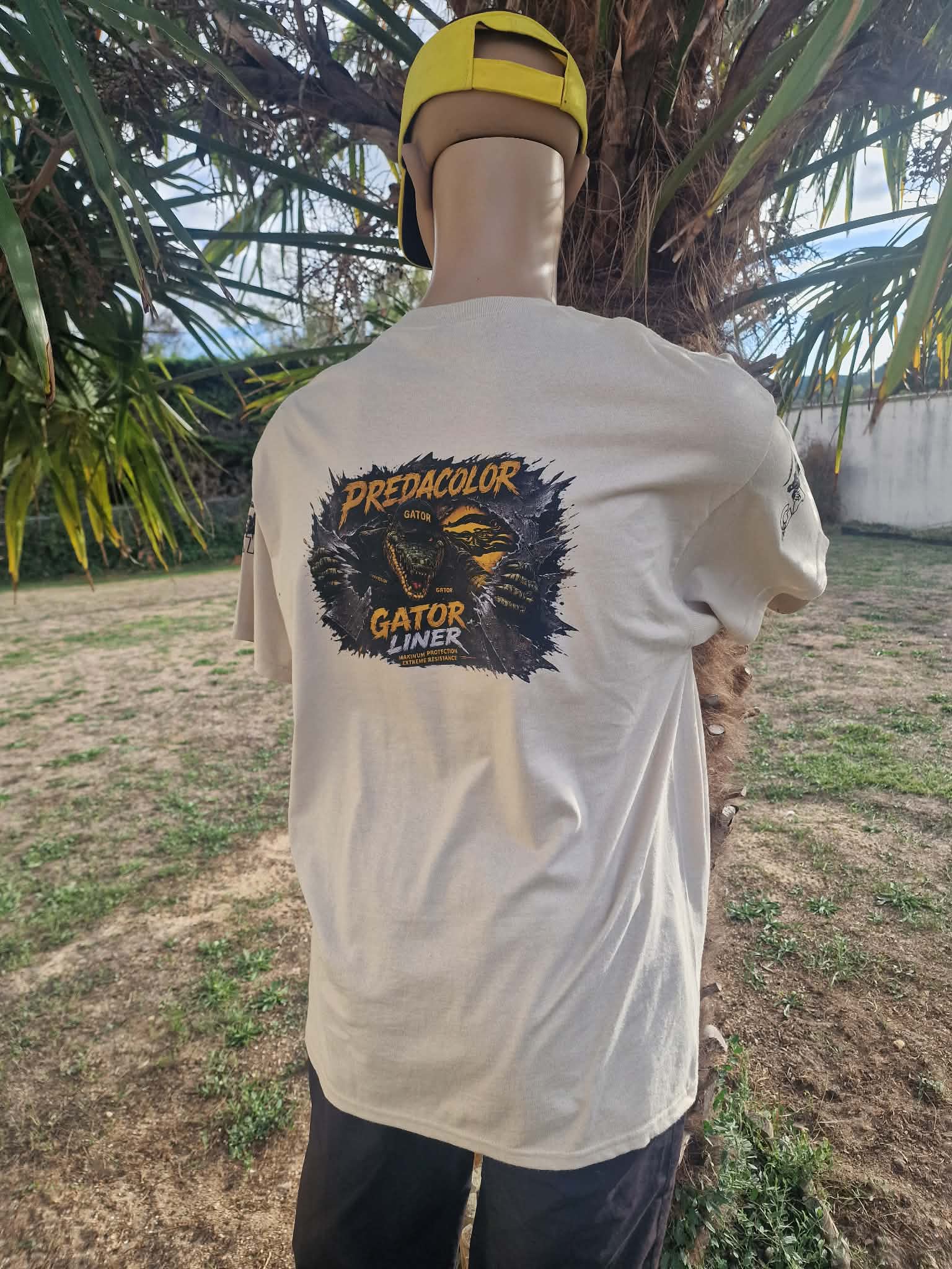 1 2 DOS - PredaColor Person seen from the back outdoors, wearing a light T-shirt with a large graphic that says 'Predacolor Gator Liner' and a yellow cap; palm trees and a fence in the background.
