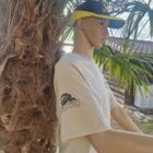 BEIGE DE COTE 1 - PredaColor Life-size mannequin wearing a beige T-shirt and a black-yellow visor hat, leaning against a palm tree outdoors with a house in the background.