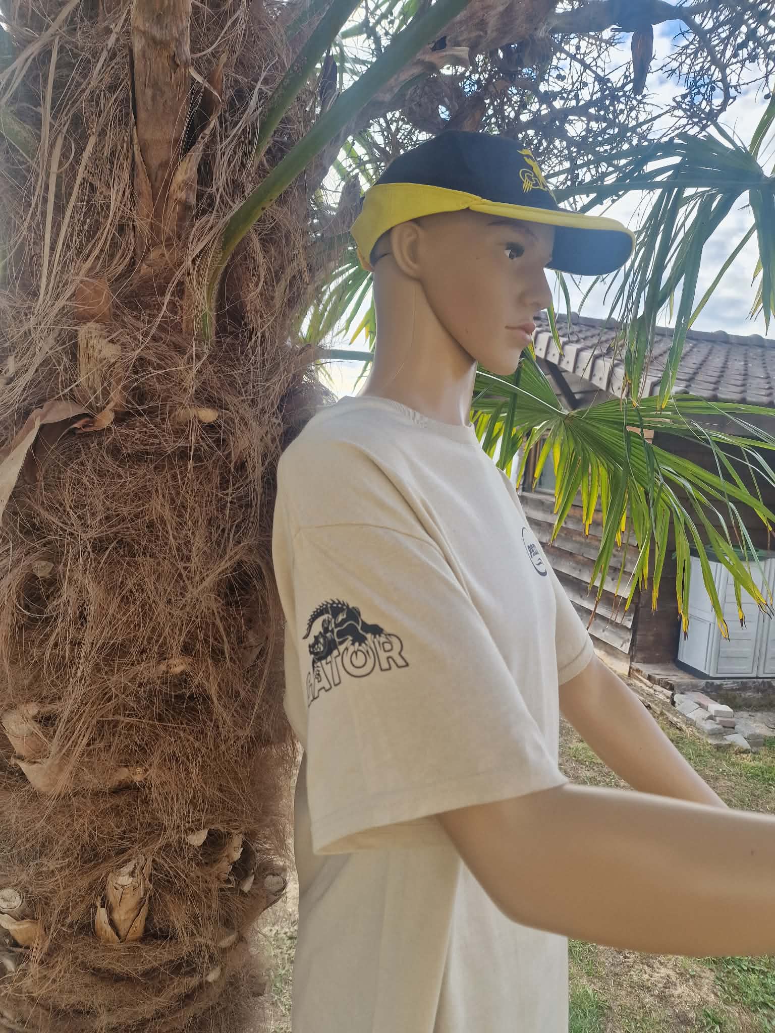 BEIGE DE COTE 1 - PredaColor Life-size mannequin wearing a beige T-shirt and a black-yellow visor hat, leaning against a palm tree outdoors with a house in the background.