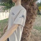 BEIGE DE COTE 2 - PredaColor Side-profile mannequin wearing a light gray T-shirt with logos and a yellow cap, beside a palm tree outdoors.