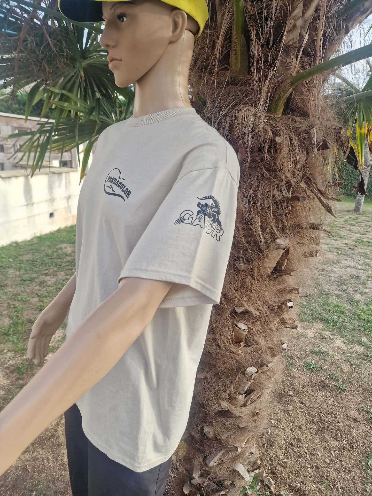 BEIGE DE COTE 2 - PredaColor Side-profile mannequin wearing a light gray T-shirt with logos and a yellow cap, beside a palm tree outdoors.