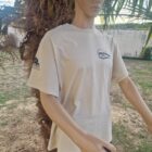 BEIGE DE FACE 2 - PredaColor Mannequin torso wearing a beige T-shirt with a circular logo on the chest, standing beside a palm tree outdoors.