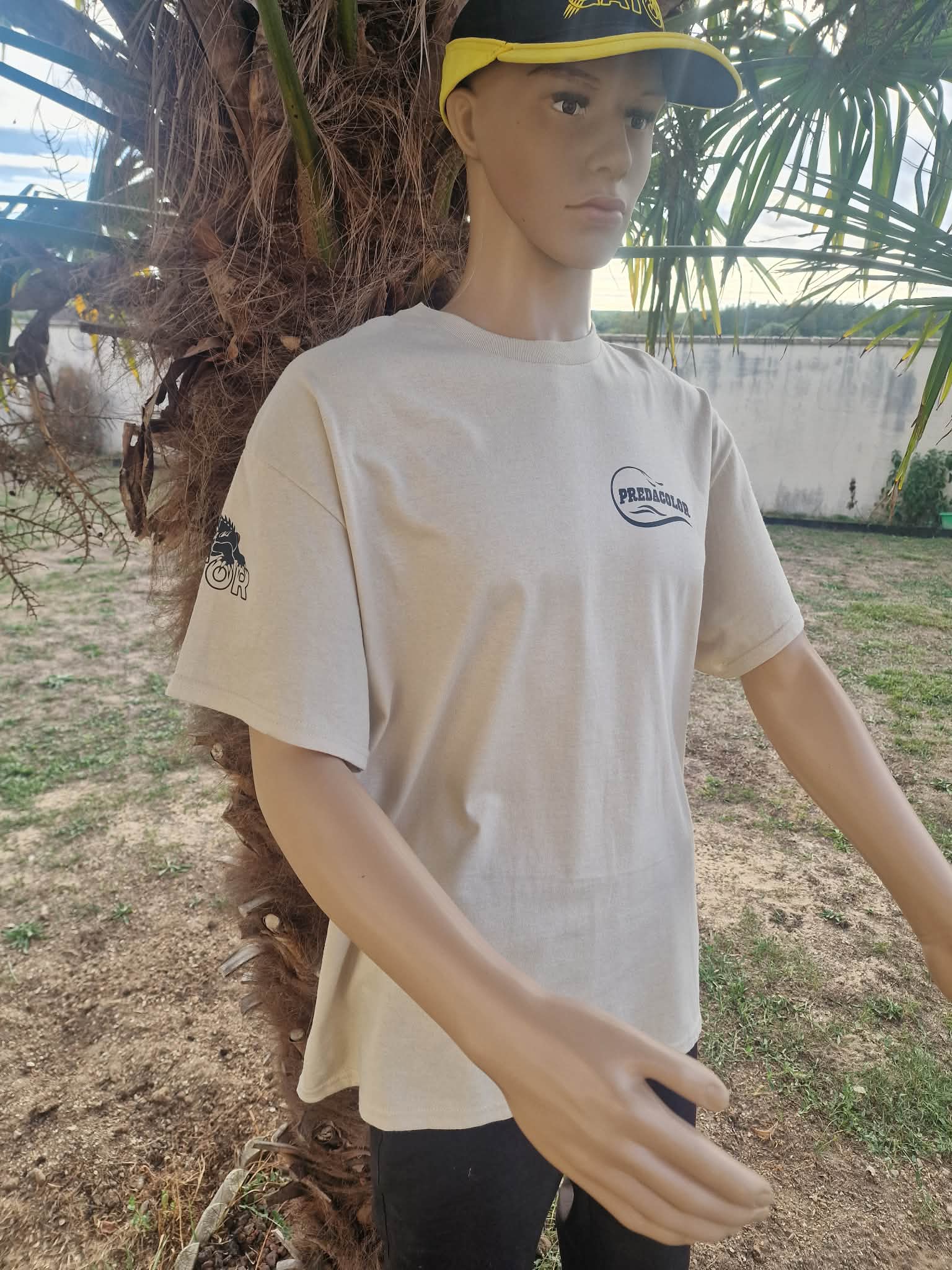 BEIGE DE FACE 2 - PredaColor Mannequin torso wearing a beige T-shirt with a circular logo on the chest, standing beside a palm tree outdoors.