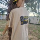 2 2 DOS - PredaColor Back view of a person wearing a beige T-shirt with a large monster-truck graphic and a yellow cap, outdoors by palm trees.
