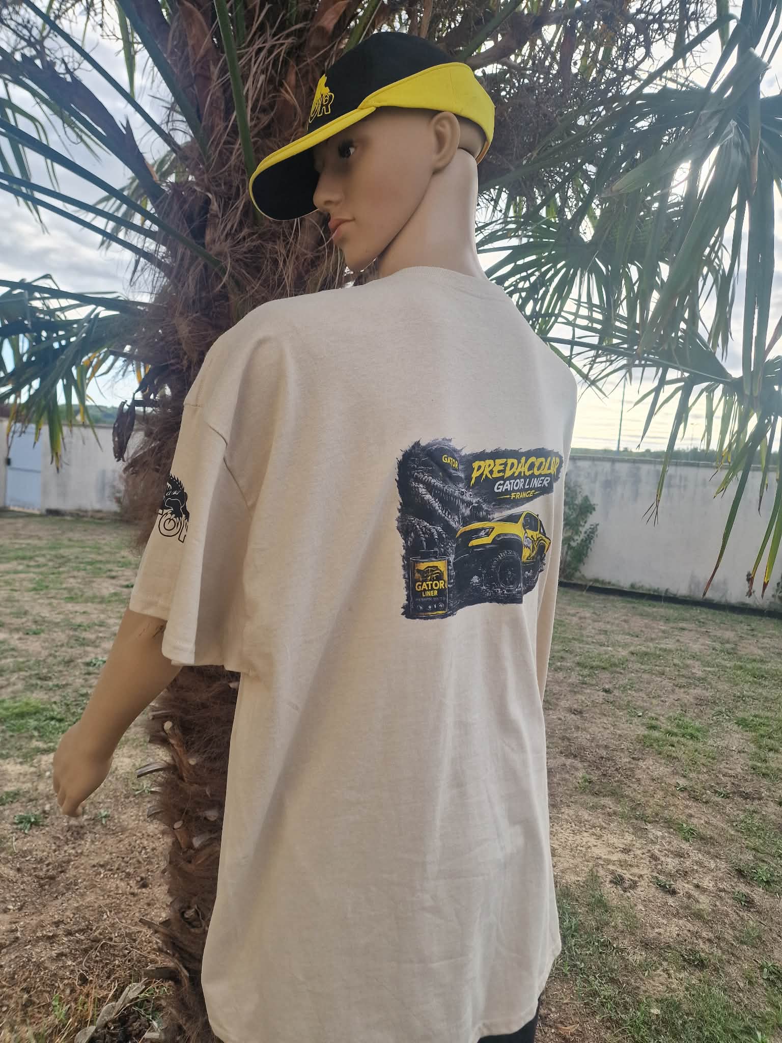 2 2 DOS - PredaColor Back view of a person wearing a beige T-shirt with a large monster-truck graphic and a yellow cap, outdoors by palm trees.