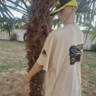 2 DOS - PredaColor Mannequin wearing a beige oversized T-shirt with a graphic on the back, standing beside a palm tree wearing a black-and-yellow cap outdoors