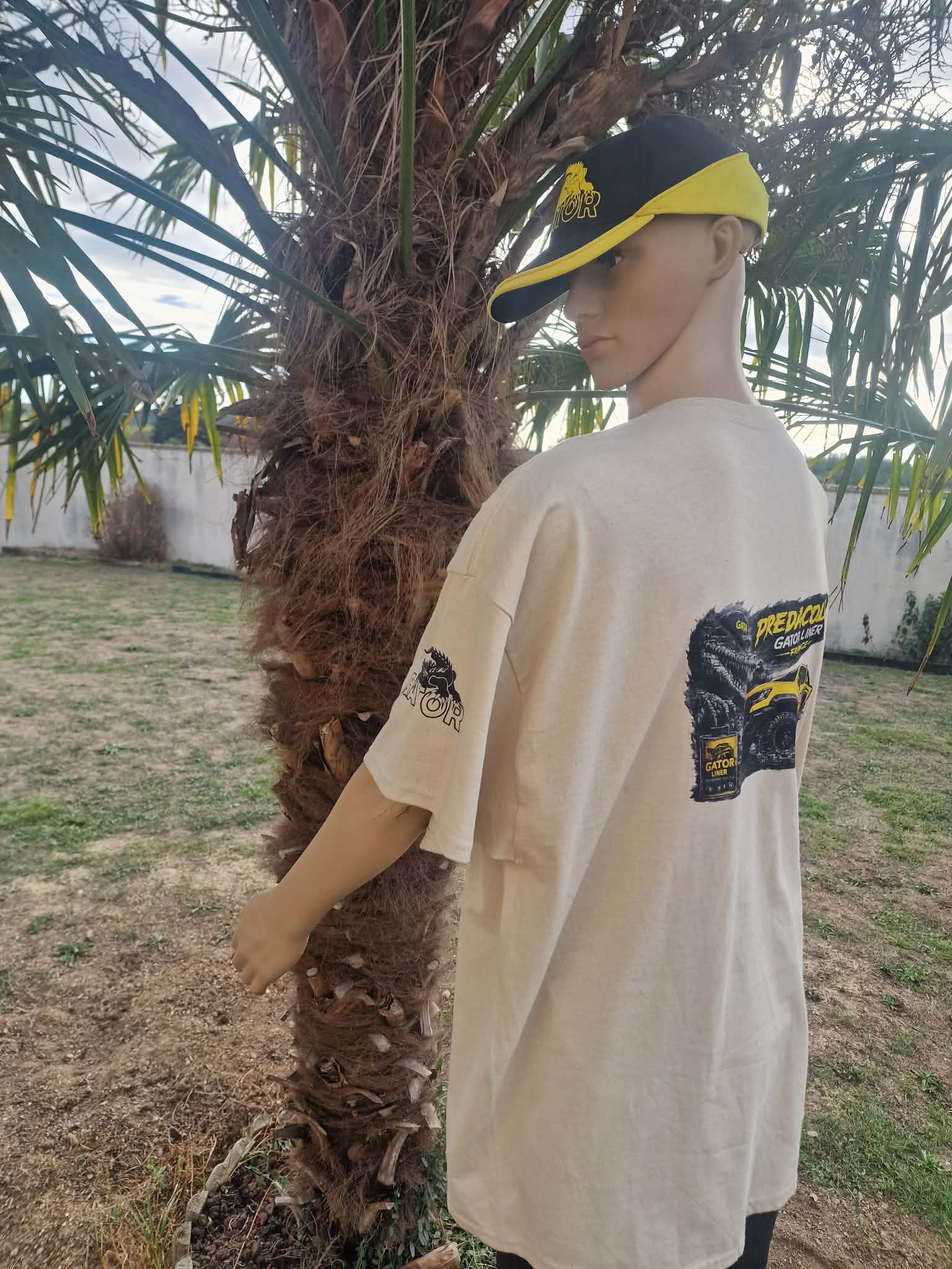 2 DOS - PredaColor Mannequin wearing a beige oversized T-shirt with a graphic on the back, standing beside a palm tree wearing a black-and-yellow cap outdoors
