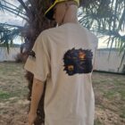 3 2 DOS - PredaColor Person seen from behind wearing a beige graphic T-shirt and a yellow cap, standing beside a palm tree in a yard.