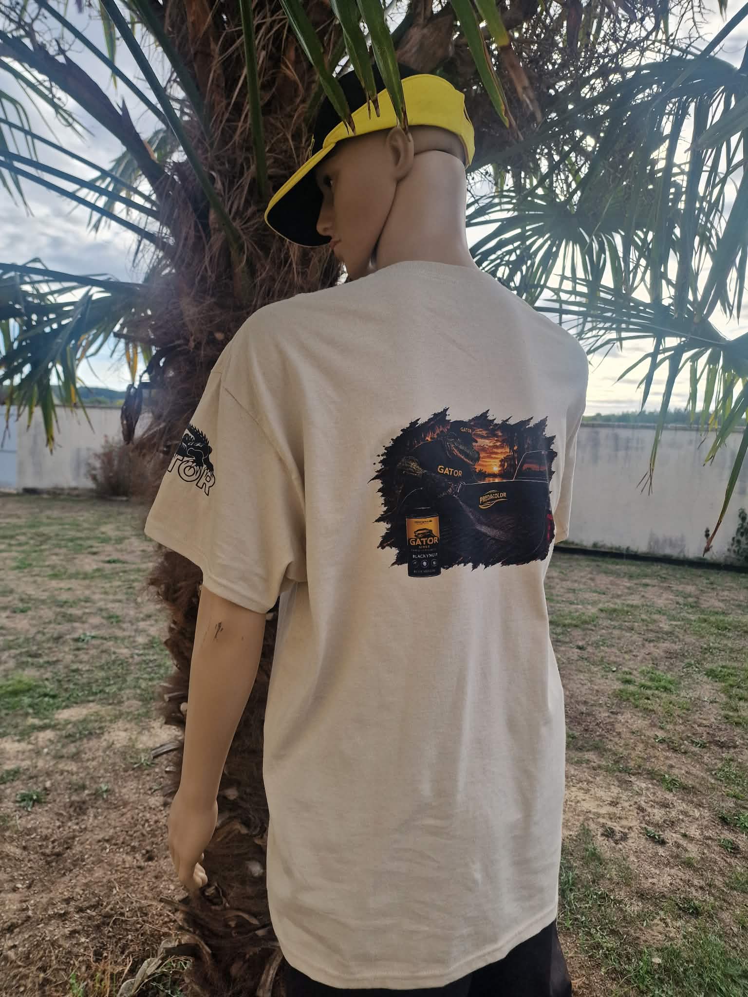 3 2 DOS - PredaColor Person seen from behind wearing a beige graphic T-shirt and a yellow cap, standing beside a palm tree in a yard.