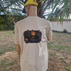 3 DOS - PredaColor Back view of a person wearing a beige graphic T-shirt and a yellow cap worn backward, standing beside a palm tree outdoors, facing away from the camera.