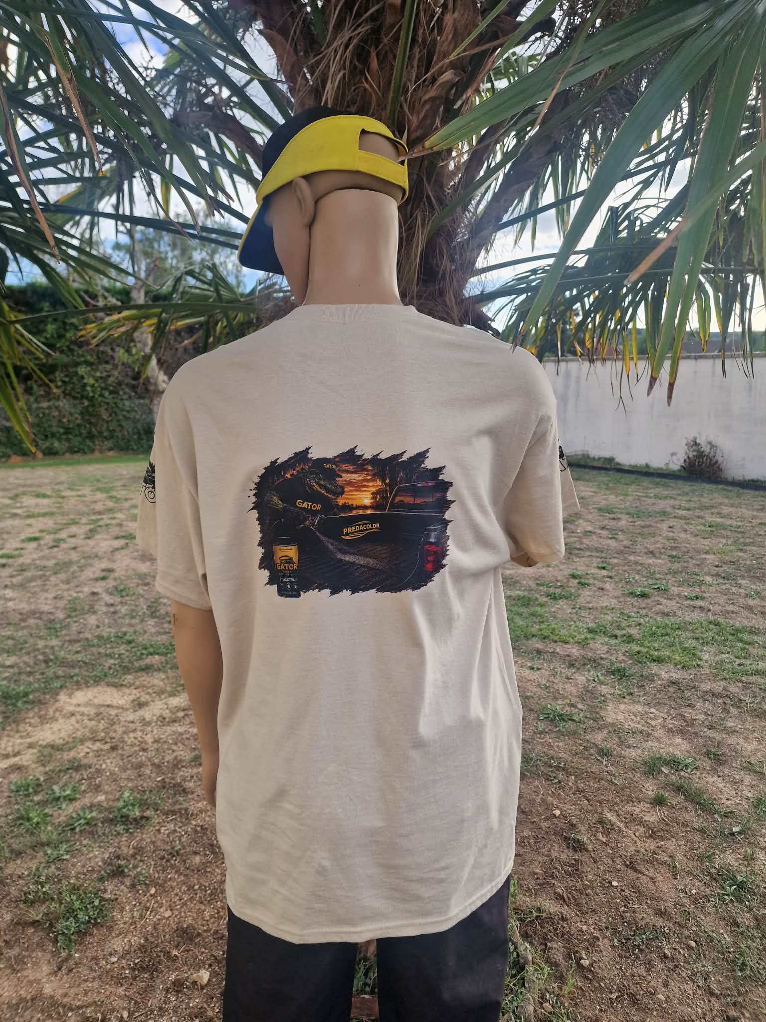 3 DOS - PredaColor Back view of a person wearing a beige graphic T-shirt and a yellow cap worn backward, standing beside a palm tree outdoors, facing away from the camera.