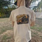4 1 DOS - PredaColor Back view of a person wearing a beige graphic T-shirt with a vintage truck design and a yellow–navy cap, outdoors by palm trees.