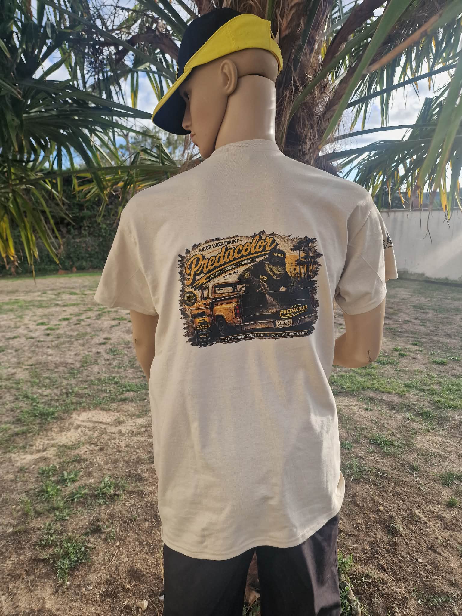 4 1 DOS - PredaColor Back view of a person wearing a beige graphic T-shirt with a vintage truck design and a yellow–navy cap, outdoors by palm trees.