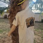 4 DOS - PredaColor Mannequin wearing a beige graphic T-shirt and a black-yellow visor cap, standing beside a palm tree outdoors.