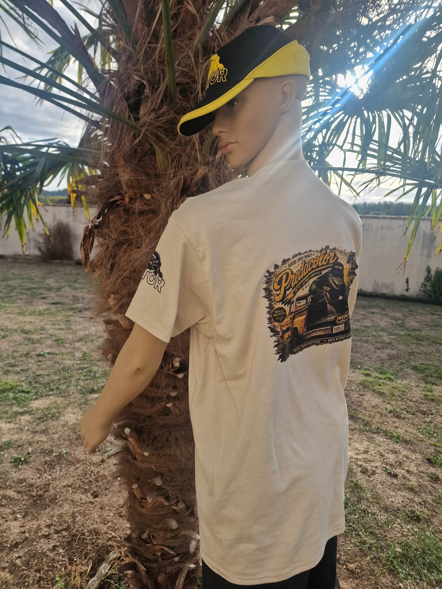 4 DOS - PredaColor Mannequin wearing a beige graphic T-shirt and a black-yellow visor cap, standing beside a palm tree outdoors.