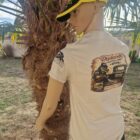 Side-view mannequin leaning against a palm tree, wearing a beige T-shirt with a large back graphic and a yellow-black cap.