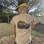Back view of a mannequin wearing a beige T-shirt with a graphic and a yellow/black cap, outdoors near a palm tree.