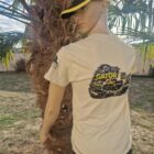 Life-size mannequin in a beige T-shirt with a graphic on the back, wearing a yellow and black cap, posed beside a palm tree in a sunny outdoor setting.
