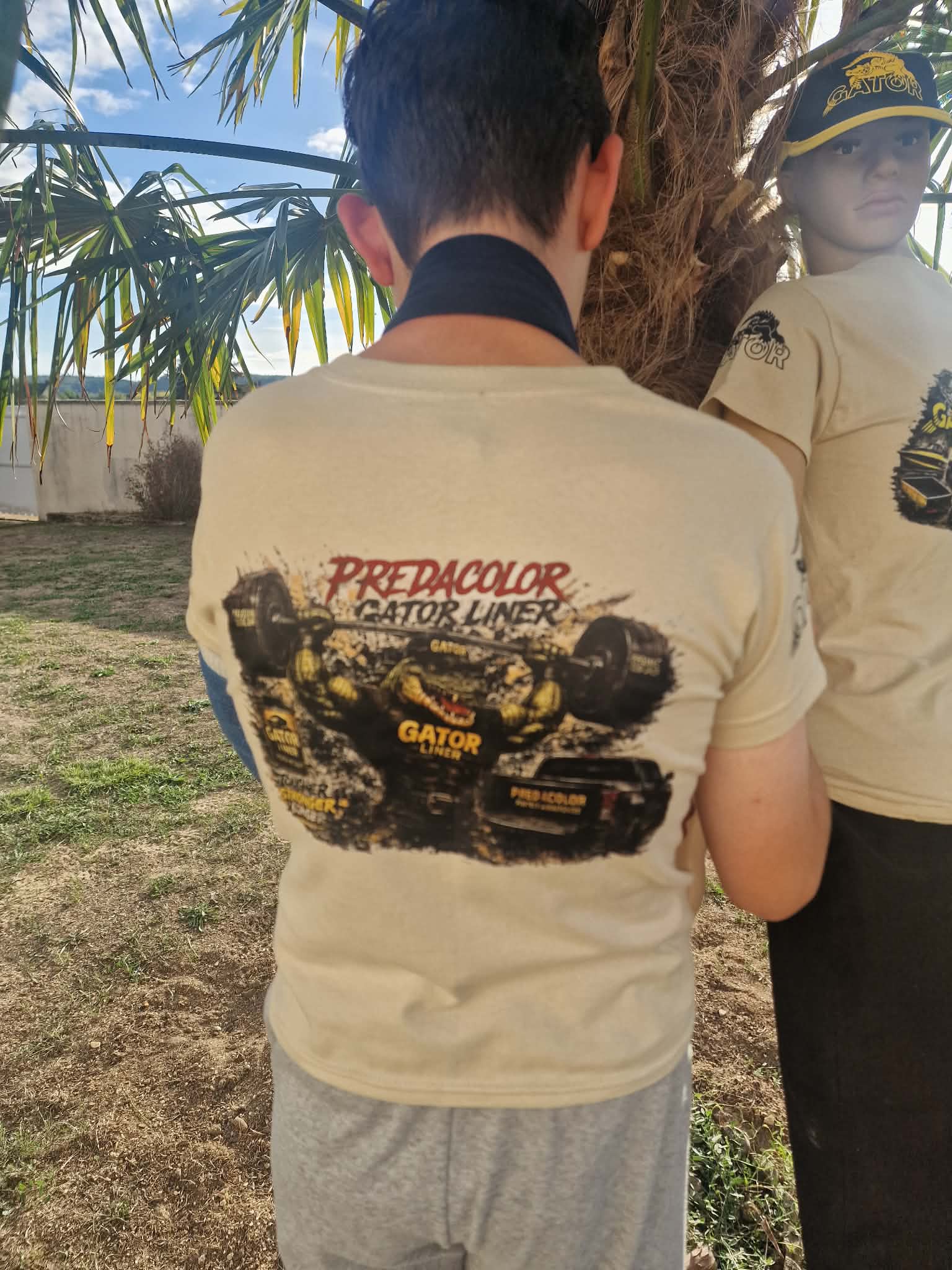 7 1 DOS - PredaColor Back view of a person in a beige T-shirt with a racing graphic on the back, standing beside a mannequin under a palm tree.