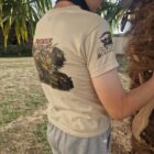 7 2 DOS - PredaColor Back view of a person in a beige T-shirt with a graphic on the back, standing near a palm tree outdoors.