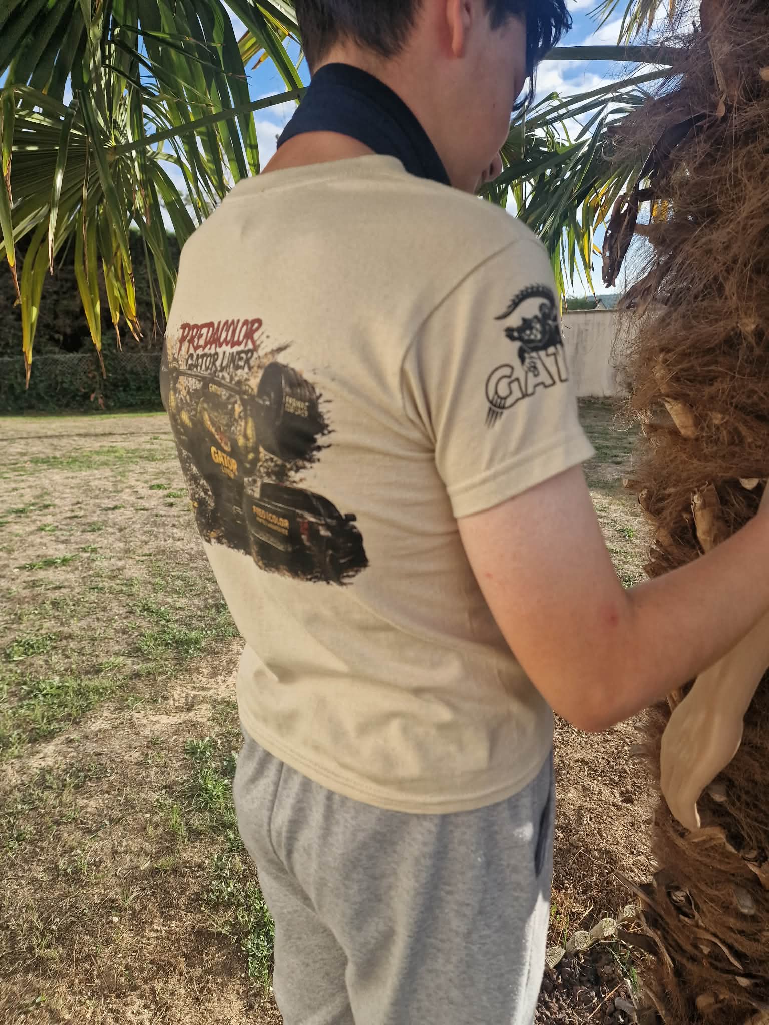 7 2 DOS - PredaColor Back view of a person in a beige T-shirt with a graphic on the back, standing near a palm tree outdoors.