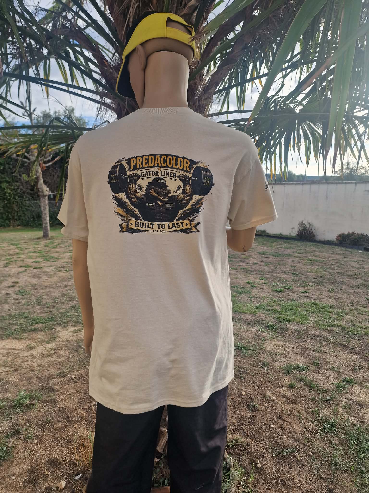 8 1 DOS - PredaColor Person standing with back to camera, wearing a beige graphic T-shirt and a yellow cap, outdoors near a palm tree.