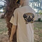 8 2 DOS - PredaColor Back view of a person/mannequin wearing a beige T-shirt with a graphic on the back, standing beside a palm tree outdoors, wearing a yellow cap.