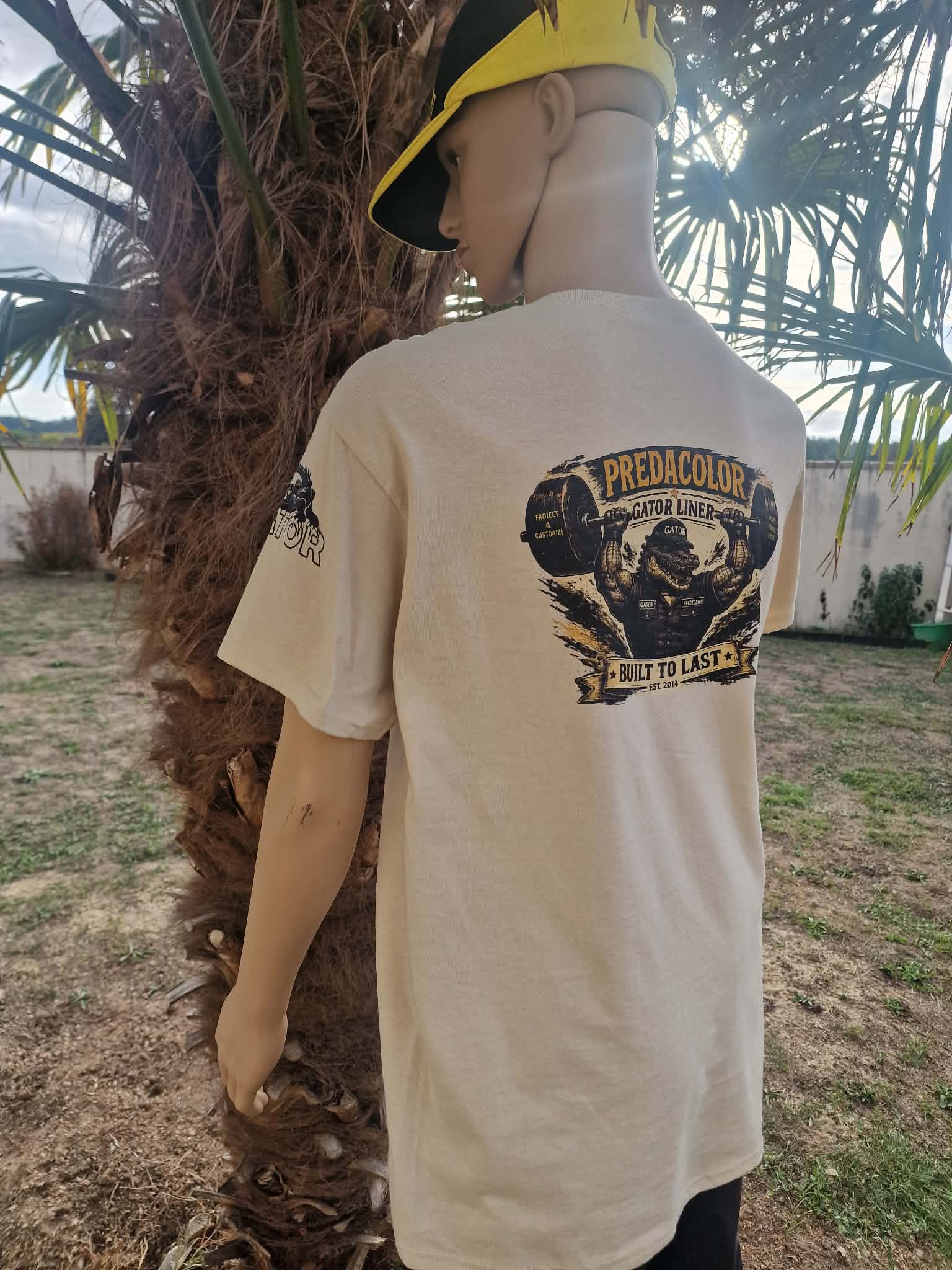 8 2 DOS - PredaColor Back view of a person/mannequin wearing a beige T-shirt with a graphic on the back, standing beside a palm tree outdoors, wearing a yellow cap.